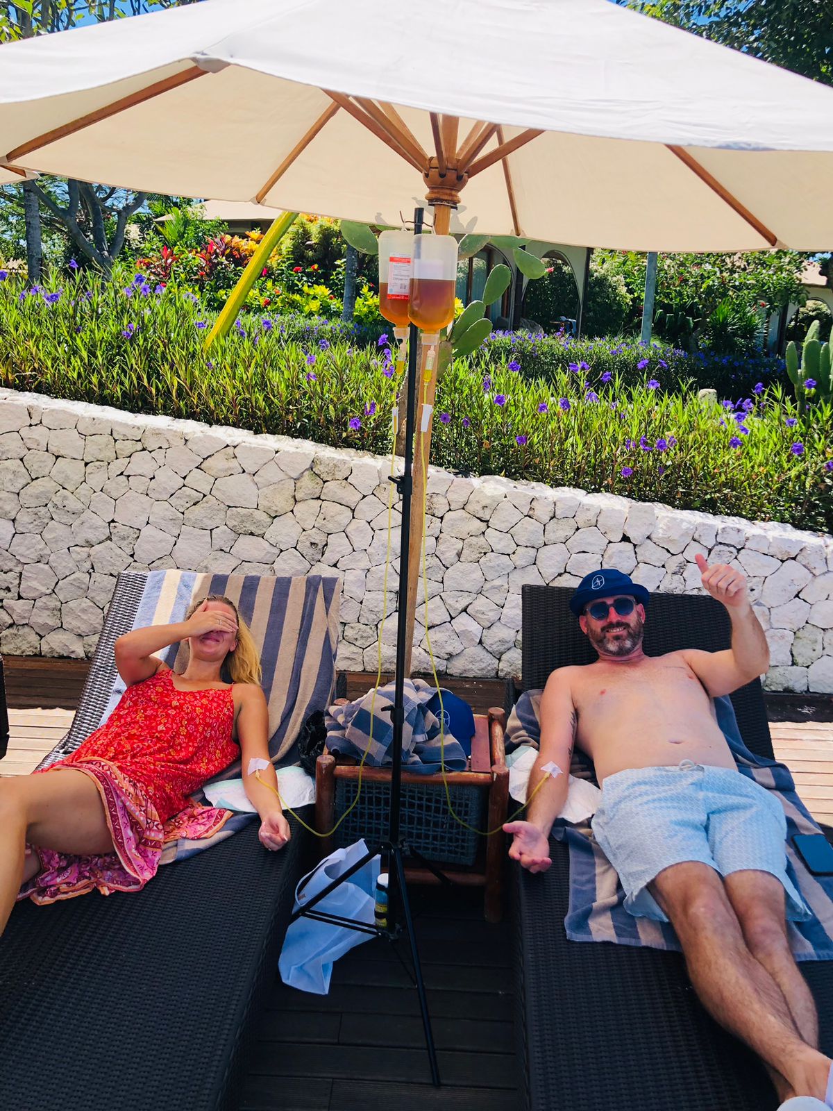 Couple relaxing on sun loungers under an umbrella receiving mobile IV drip therapy in Bali.